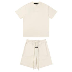 May include: A cream-colored short-sleeve t-shirt and matching shorts set. The t-shirt features a black rectangular label at the neckline. The shorts have an elastic waistband with long cream drawstrings and a black rectangular label at the center.