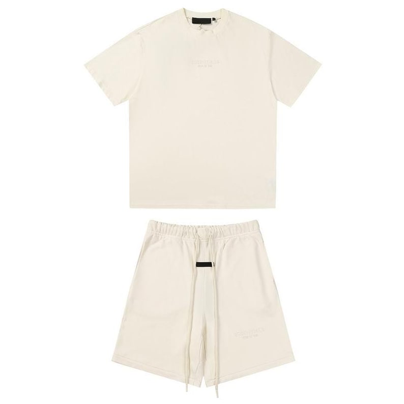 May include: A cream-colored short-sleeve t-shirt and matching shorts set. The t-shirt features a black rectangular label at the neckline. The shorts have an elastic waistband with long cream drawstrings and a black rectangular label at the center.