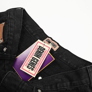 May include: Black denim jeans featuring a tan leather patch on the pocket and a rectangular tag attached to the waistband. The tag displays the text DENIM GENES, LUNDUN.INGERLAND.2018, and CORTEIZRULESTHEWORLD in black, white, and purple colors.
