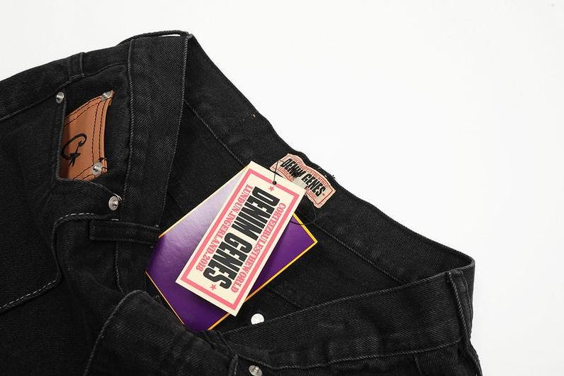 May include: Black denim jeans featuring a tan leather patch on the pocket and a rectangular tag attached to the waistband. The tag displays the text DENIM GENES, LUNDUN.INGERLAND.2018, and CORTEIZRULESTHEWORLD in black, white, and purple colors.