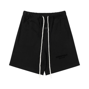 May include: Black shorts featuring an elastic waistband with long white drawstring cords. The front displays the text ESSENTIALS FEAR OF GOD in a tonal black print, and a small rectangular black patch is centered just below the waistband.