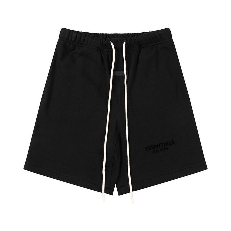 May include: Black shorts featuring an elastic waistband with long white drawstring cords. The front displays the text ESSENTIALS FEAR OF GOD in a tonal black print, and a small rectangular black patch is centered just below the waistband.
