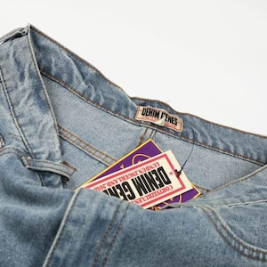 Peut inclure: Un jean en denim bleu clair avec une &eacute;tiquette de marque Denim Genes sur la ceinture. Une &eacute;tiquette volante rectangulaire est attach&eacute;e, affichant le texte Denim Genes, Lundun Ingerland 2010 et Corteizrules en blanc, rouge et violet sur la carte.