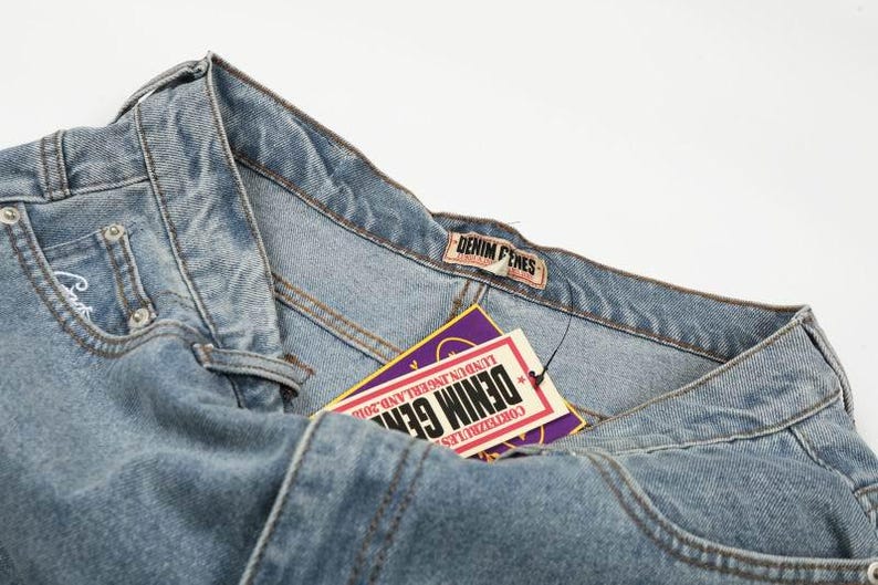 Peut inclure: Un jean en denim bleu clair avec une &eacute;tiquette de marque Denim Genes sur la ceinture. Une &eacute;tiquette volante rectangulaire est attach&eacute;e, affichant le texte Denim Genes, Lundun Ingerland 2010 et Corteizrules en blanc, rouge et violet sur la carte.