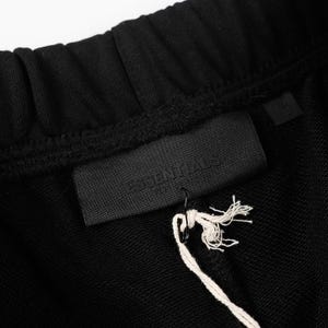 May include: Black sweatpants featuring an elasticized waistband and a rectangular black fabric patch sewn onto the front. The patch displays the embossed text ESSENTIALS FEAR OF GOD. A white string tag is attached to the patch with a black pin.