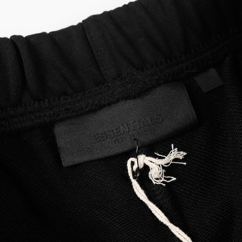May include: Black sweatpants featuring an elasticized waistband and a rectangular black fabric patch sewn onto the front. The patch displays the embossed text ESSENTIALS FEAR OF GOD. A white string tag is attached to the patch with a black pin.
