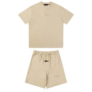 May include: A beige short-sleeve t-shirt and matching shorts set. The t-shirt features a crew neckline and the text ESSENTIALS FEAR OF GOD printed on the chest. The shorts have an elastic waistband with long white drawstrings and the same text.