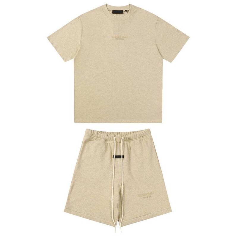 May include: A beige short-sleeve t-shirt and matching shorts set. The t-shirt features a crew neckline and the text ESSENTIALS FEAR OF GOD printed on the chest. The shorts have an elastic waistband with long white drawstrings and the same text.