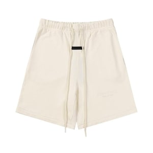 May include: Off-white sweat shorts featuring an elasticized waistband with long, matching drawstrings. A small black rectangular patch is centered below the waistband. The word Essentials is embossed on the lower left leg in a tonal cream color.