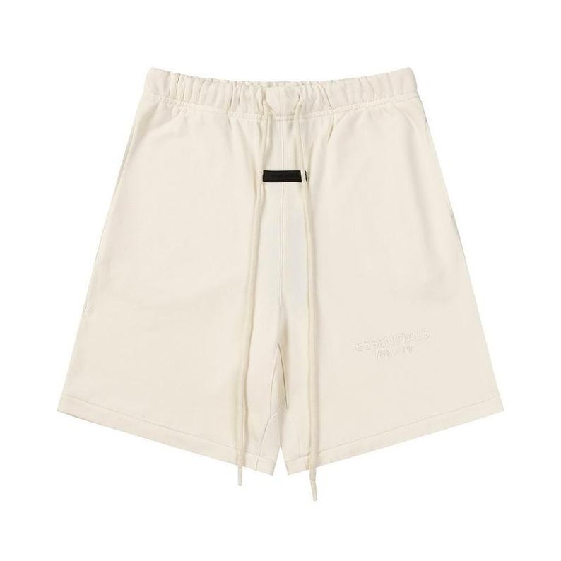 May include: Off-white sweat shorts featuring an elasticized waistband with long, matching drawstrings. A small black rectangular patch is centered below the waistband. The word Essentials is embossed on the lower left leg in a tonal cream color.