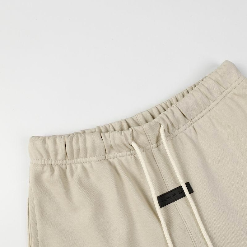 May include: Beige sweatpants featuring an elasticized waistband with a drawstring closure. A small, rectangular black patch with white text is centered below the waistband, positioned between the two white drawstring cords.