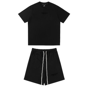 May include: A black short-sleeve t-shirt and matching black shorts set. The t-shirt features the word ESSENTIALS printed on the chest. The shorts have an elastic waistband with long white drawstrings and the word ESSENTIALS printed on the leg.