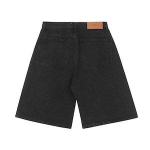 May include: Black denim shorts featuring a high-waisted design with two back pockets and a tan leather-colored rectangular patch on the waistband. The shorts have a straight-leg cut and a classic five-pocket style construction in dark fabric.