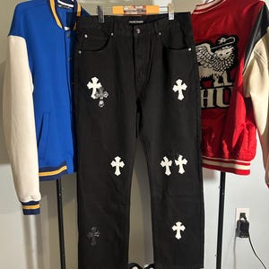 May include: Black denim jeans with white cross patches and a distressed knee. A blue and white varsity jacket and a red and white jacket hang on a clothing rack. Red and white sneakers are at the bottom.