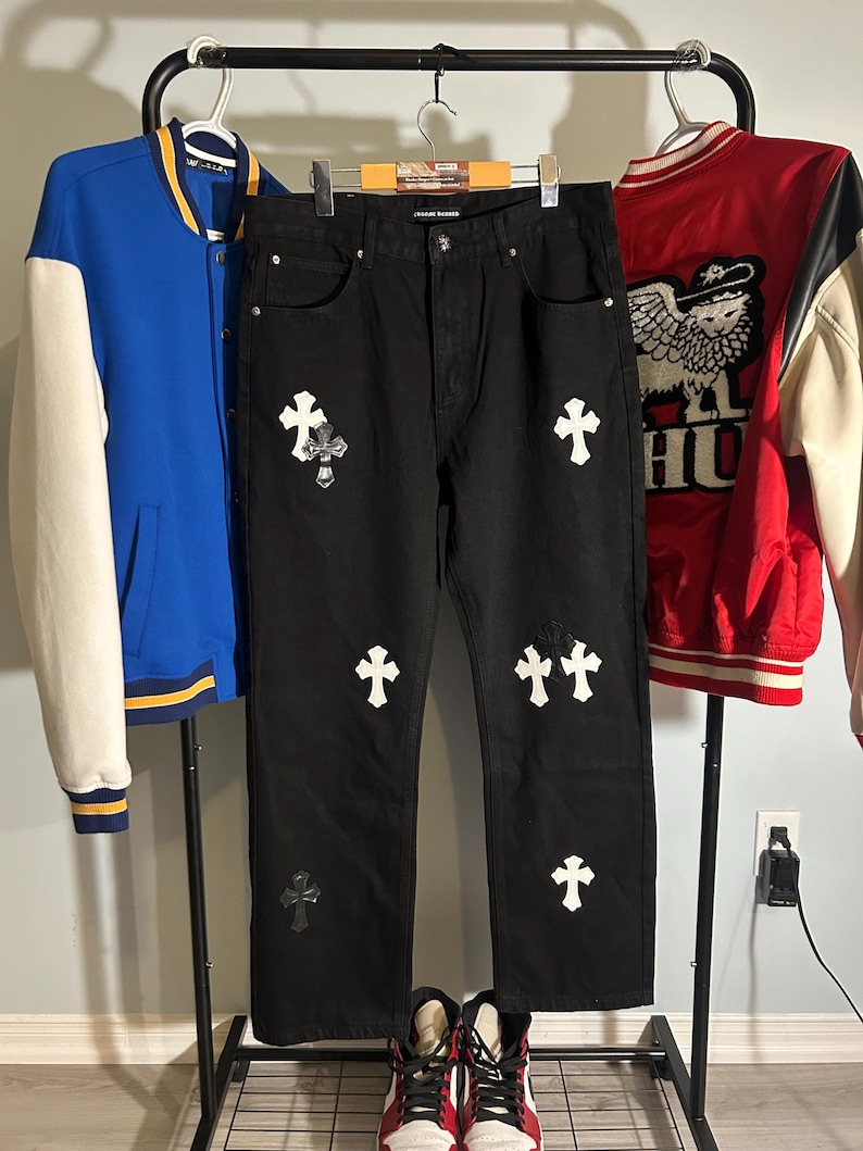 May include: Black denim jeans with white cross patches and a distressed knee. A blue and white varsity jacket and a red and white jacket hang on a clothing rack. Red and white sneakers are at the bottom.