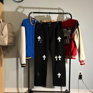 May include: A black pair of jeans with white cross appliques, flanked by a blue and white varsity jacket and a red and white varsity jacket. The clothing is displayed on a rolling rack.