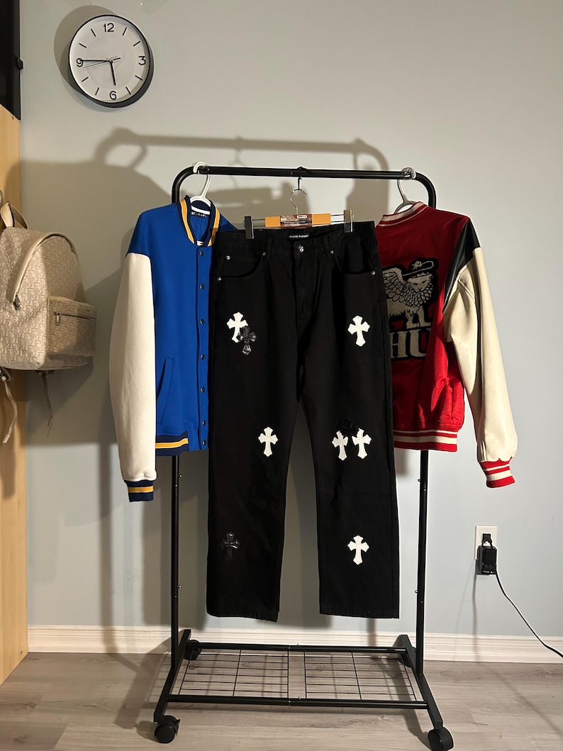 May include: A black pair of jeans with white cross appliques, flanked by a blue and white varsity jacket and a red and white varsity jacket. The clothing is displayed on a rolling rack.