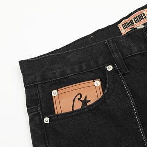 May include: Black denim jeans featuring a tan leather patch on the front pocket with a black star and letter C logo. The waistband includes a tan label with the text DENIM GENES. The jeans have silver-toned metal rivets and visible stitching.