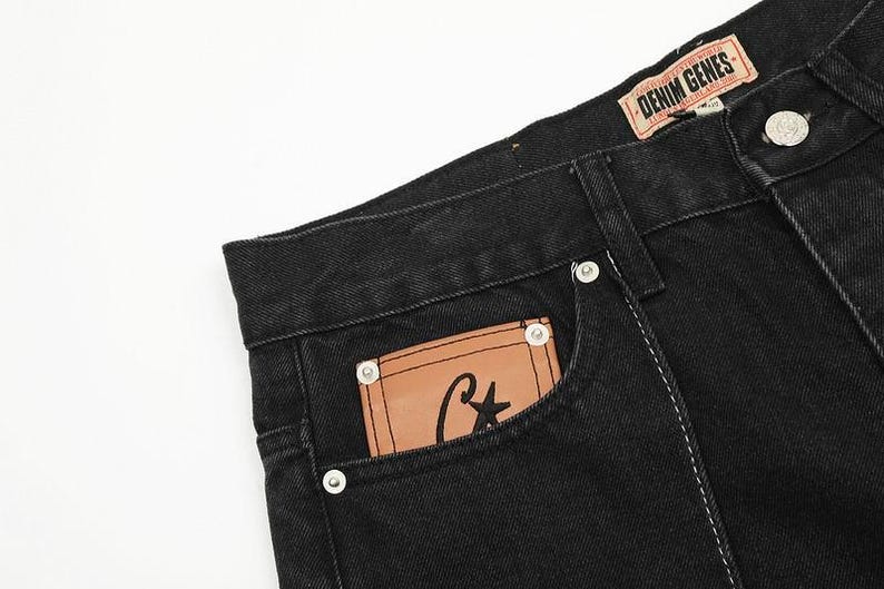 May include: Black denim jeans featuring a tan leather patch on the front pocket with a black star and letter C logo. The waistband includes a tan label with the text DENIM GENES. The jeans have silver-toned metal rivets and visible stitching.