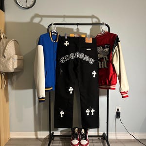 May include: A black rolling clothing rack displays a blue and white letterman jacket, black jeans with white cross and text details, and a red and white jacket. A pair of red and white high-top sneakers sit below the jeans.