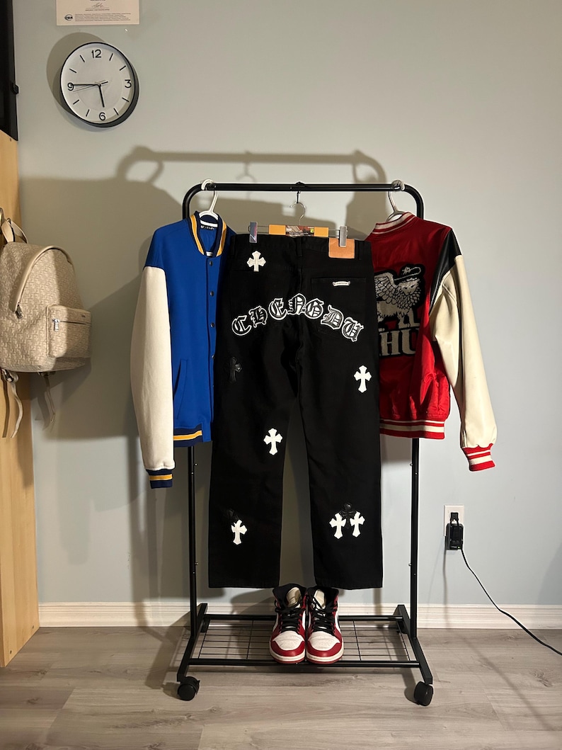 May include: A black rolling clothing rack displays a blue and white letterman jacket, black jeans with white cross and text details, and a red and white jacket. A pair of red and white high-top sneakers sit below the jeans.