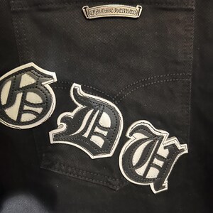 May include: Close-up of black denim jeans with three white and black leather patches. The patches feature stylized letters. A silver metal tag with the text "CHROME HEARTS" is above the back pocket.