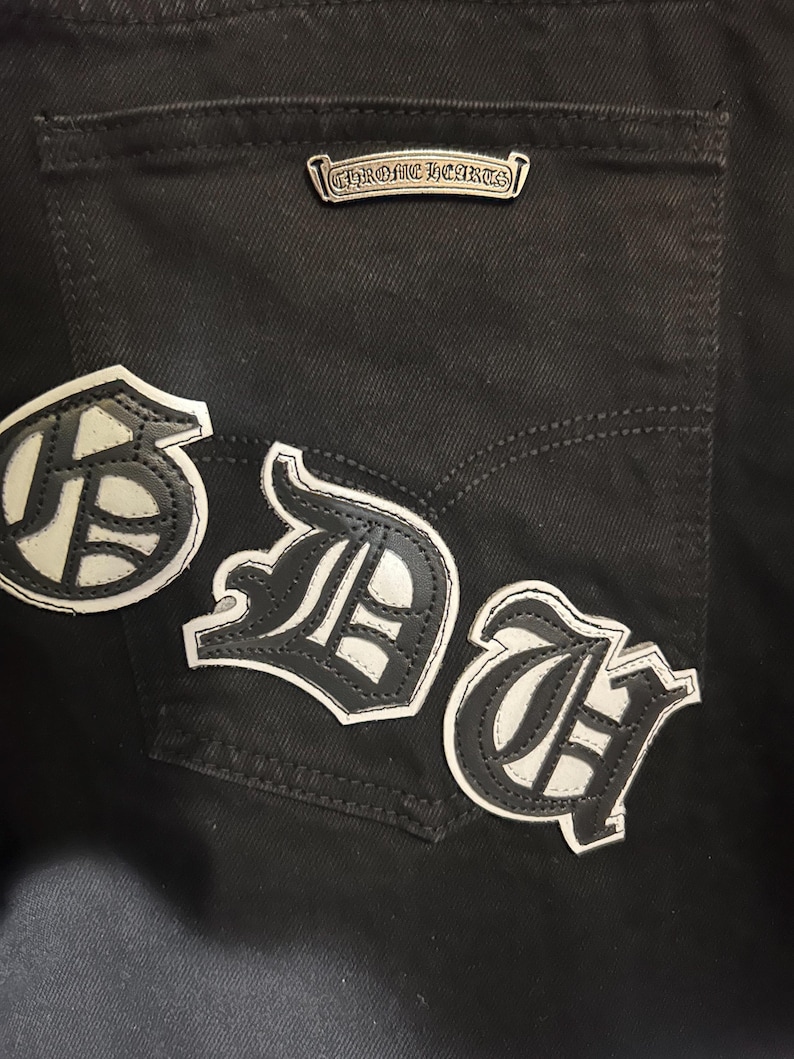 May include: Close-up of black denim jeans with three white and black leather patches. The patches feature stylized letters. A silver metal tag with the text "CHROME HEARTS" is above the back pocket.