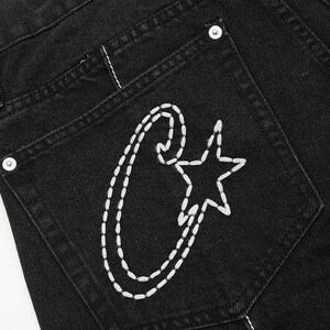 May include: Black denim jeans featuring a back pocket with white hand-stitched embroidery. The design consists of a large, stylized letter C combined with a five-pointed star. A silver-toned metal rivet is visible at the top corner of the pocket.