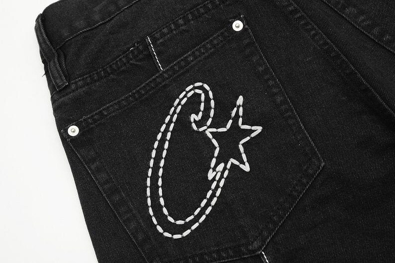 May include: Black denim jeans featuring a back pocket with white hand-stitched embroidery. The design consists of a large, stylized letter C combined with a five-pointed star. A silver-toned metal rivet is visible at the top corner of the pocket.