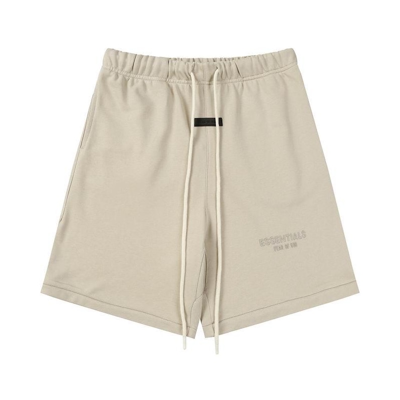 May include: Beige shorts featuring an elastic waistband with long, off-white drawstring cords. A small black rectangular patch is centered below the waistband, and the word ESSENTIALS is printed in a tonal beige font on the lower left leg.