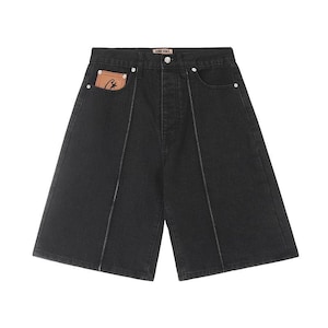 May include: Black denim shorts featuring a high-waisted design with a button fly closure. The shorts include vertical white contrast stitching down the front, silver-tone rivets, and a tan leather-like patch with a black logo inside the pocket.