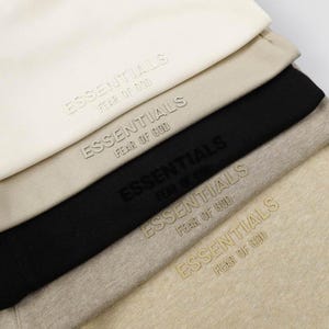 May include: A stack of five folded garments in shades of cream, beige, black, and light brown. Each piece features the embossed text ESSENTIALS FEAR OF GOD on the front, with the lettering color-matched to the fabric of each individual item.