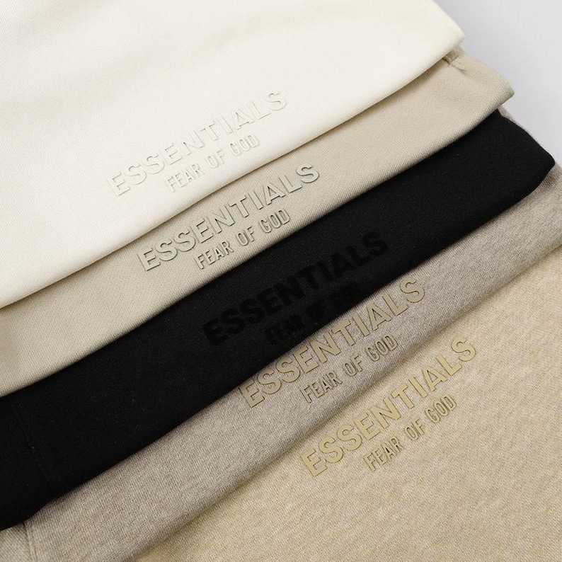 May include: A stack of five folded garments in shades of cream, beige, black, and light brown. Each piece features the embossed text ESSENTIALS FEAR OF GOD on the front, with the lettering color-matched to the fabric of each individual item.