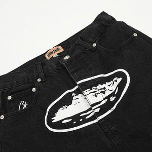 May include: Black denim jeans featuring a large white oval graphic print on the front. The waistband includes a tan label with the text DENIM GENES and silver-toned metal rivets. A small white star logo is embroidered on the front pocket area.
