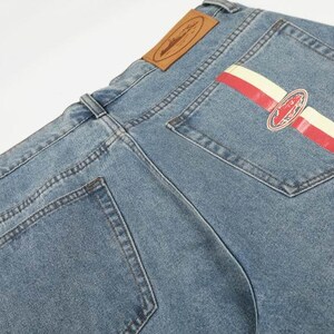 Peut inclure: Jean en denim bleu clair avec une &eacute;tiquette en cuir beige sur la ceinture arri&egrave;re. La poche arri&egrave;re droite pr&eacute;sente un motif &agrave; rayures horizontales rouges et cr&egrave;me avec un embl&egrave;me ovale central contenant des d&eacute;tails graphiques et du texte.