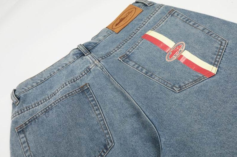 Peut inclure: Jean en denim bleu clair avec une &eacute;tiquette en cuir beige sur la ceinture arri&egrave;re. La poche arri&egrave;re droite pr&eacute;sente un motif &agrave; rayures horizontales rouges et cr&egrave;me avec un embl&egrave;me ovale central contenant des d&eacute;tails graphiques et du texte.