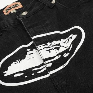 May include: Black denim jeans featuring a large white graphic print of an oval shape containing a stylized landscape. The waistband includes a tan leather-colored patch with the text DENIM GENES in black, silver metal rivets, and a button closure.