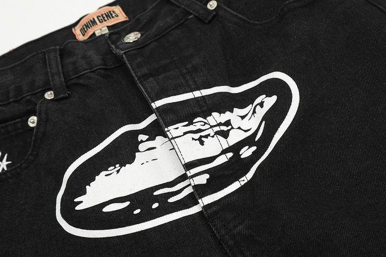 May include: Black denim jeans featuring a large white graphic print of an oval shape containing a stylized landscape. The waistband includes a tan leather-colored patch with the text DENIM GENES in black, silver metal rivets, and a button closure.