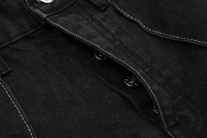 May include: A pair of black denim jeans featuring a button-fly closure with metallic silver buttons. The fabric has a visible twill texture and is accented with contrasting white stitching along the seams and the placket of the fly.