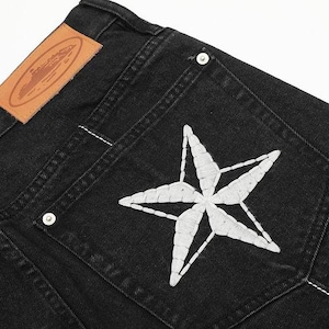 May include: Black denim jeans featuring a large white embroidered star on the back pocket, silver-toned metal rivets, and a tan leather patch on the waistband with an embossed oval logo design and visible white stitching along the seams.