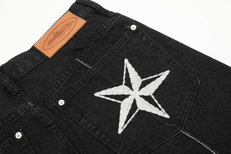 May include: Black denim jeans featuring a large white embroidered star on the back pocket, silver-toned metal rivets, and a tan leather patch on the waistband with an embossed oval logo design and visible white stitching along the seams.