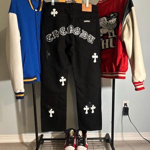May include: A display of streetwear clothing including a blue and white varsity jacket, black jeans with white cross and lettering details, and a red and white varsity jacket. A pair of red and white high-top sneakers are at the bottom.