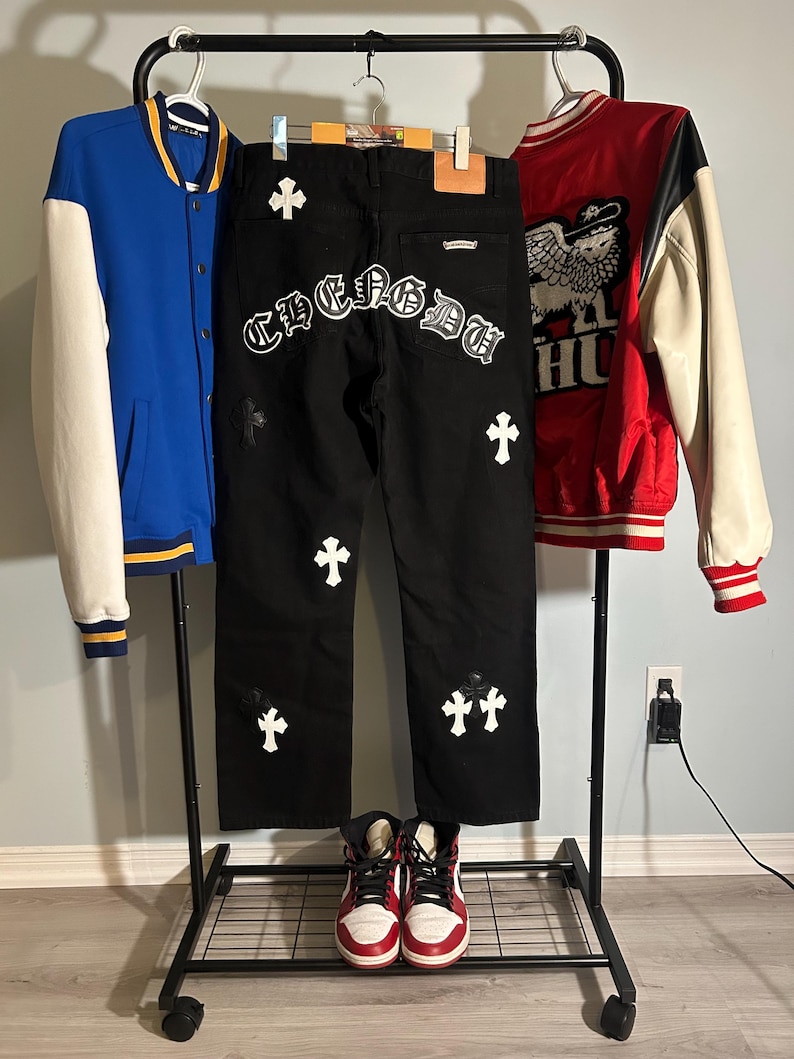 May include: A display of streetwear clothing including a blue and white varsity jacket, black jeans with white cross and lettering details, and a red and white varsity jacket. A pair of red and white high-top sneakers are at the bottom.