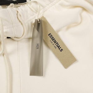 May include: Off-white sweatpants featuring an elastic waistband with a matching drawstring. Two rectangular tags are attached: one is a translucent grey plastic tag labeled MEN, and the other is a tan paper tag printed with ESSENTIALS FEAR OF GOD.
