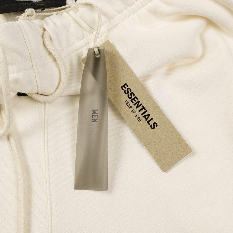 May include: Off-white sweatpants featuring an elastic waistband with a matching drawstring. Two rectangular tags are attached: one is a translucent grey plastic tag labeled MEN, and the other is a tan paper tag printed with ESSENTIALS FEAR OF GOD.