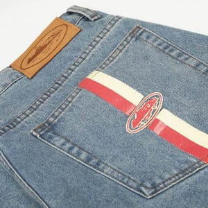 Peut inclure: Jean en denim bleu clair avec une &eacute;tiquette en cuir beige sur la ceinture et une poche arri&egrave;re orn&eacute;e d'une bande horizontale rouge et cr&egrave;me, ainsi qu'un embl&egrave;me ovale central repr&eacute;sentant la silhouette rouge d'un navire.