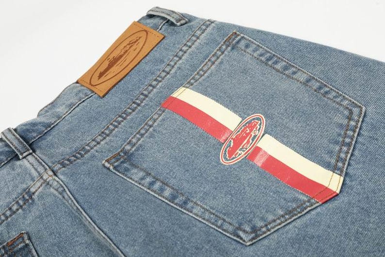 Peut inclure: Jean en denim bleu clair avec une &eacute;tiquette en cuir beige sur la ceinture et une poche arri&egrave;re orn&eacute;e d'une bande horizontale rouge et cr&egrave;me, ainsi qu'un embl&egrave;me ovale central repr&eacute;sentant la silhouette rouge d'un navire.