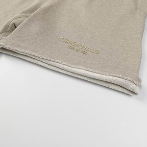 May include: Beige sweat shorts featuring a raw, unfinished hemline. The fabric has a textured, heathered appearance. Embroidered on the lower front in a matching beige tone is the text ESSENTIALS FEAR OF GOD in a clean, sans-serif font.