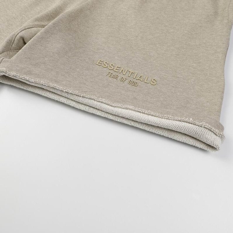 May include: Beige sweat shorts featuring a raw, unfinished hemline. The fabric has a textured, heathered appearance. Embroidered on the lower front in a matching beige tone is the text ESSENTIALS FEAR OF GOD in a clean, sans-serif font.