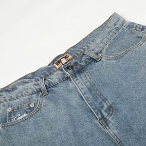 Peut inclure: Jean en denim bleu clair avec une taille haute, une fermeture &agrave; bouton et une braguette zipp&eacute;e. La ceinture comporte des passants et une petite &eacute;tiquette rectangulaire de marque Denim Bones. Une petite poche gousset pr&eacute;sente une broderie.